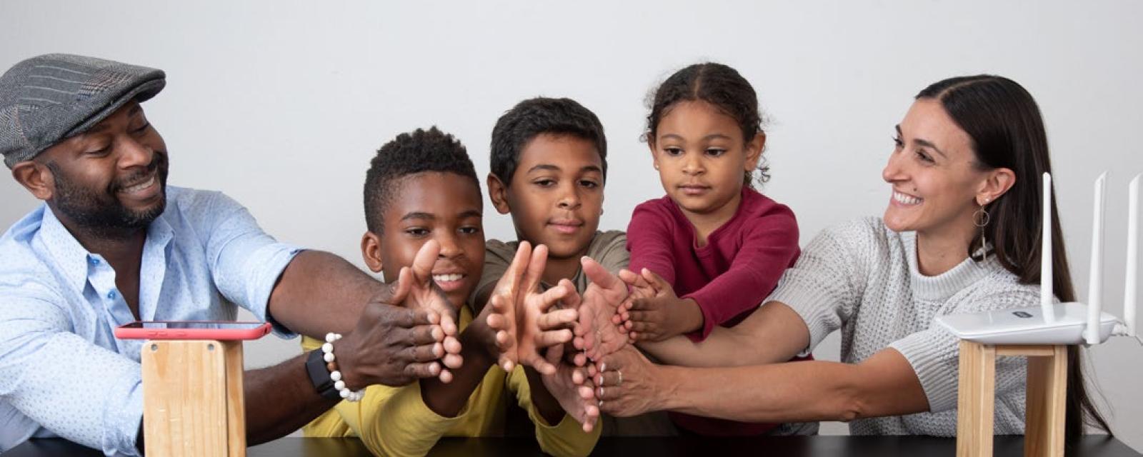 A family holds all their hands together in between a smartphone and a Wi-Fi router sitting on two blocks of wood, placed apart on a table. 